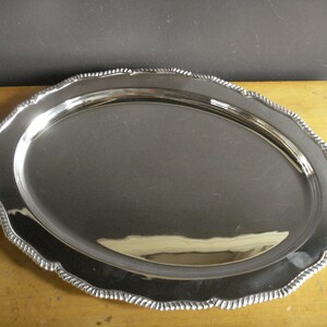 May include: A shiny, oval-shaped silver serving tray with a decorative, scalloped edge. The tray has a reflective surface, and the edge features a rope-like design. The tray is likely made of metal and is suitable for serving food or displaying items.