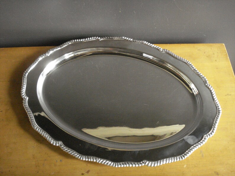May include: A shiny, oval-shaped silver serving tray with a decorative, scalloped edge. The tray has a reflective surface, and the edge features a rope-like design. The tray is likely made of metal and is suitable for serving food or displaying items.
