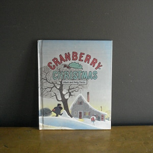 May include: A hardcover children's book titled Cranberry Christmas by Wende and Harry Devlin. The cover features a winter scene with a snow-covered house, a large bare tree, and a figure walking in the snow under a light blue sky.