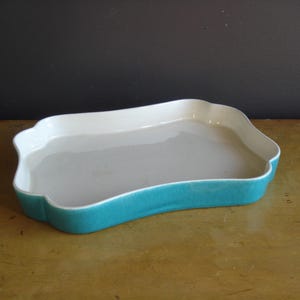May include: A rectangular ceramic baking dish with a scalloped edge. The interior is white, the base is a light gray, and the exterior is a vibrant turquoise. The dish sits on a wooden surface.