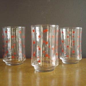 May include: Three clear drinking glasses with a red polka dot design. The glasses have a flared base and a straight, cylindrical body. The red dots are evenly spaced across the surface of each glass.