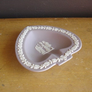 Può includere: Un piatto portaoggetti a forma di cuore di colore lavanda con un bordo floreale bianco e un cammeo bianco con tre figure al centro.