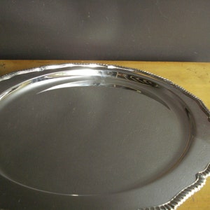 May include: A large, oval, silver-colored serving tray with a decorative, scalloped edge. The tray has a reflective surface and sits on a light-colored wooden surface. The edge has a rope-like design.