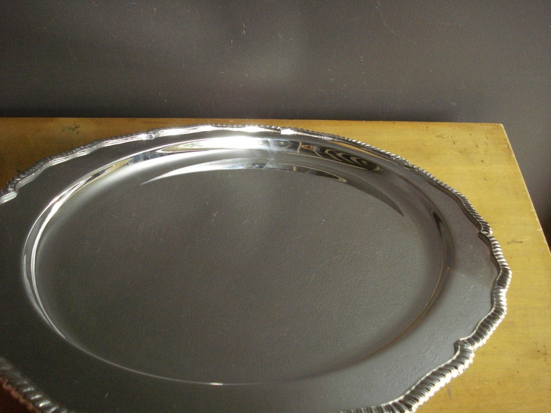 May include: A large, oval, silver-colored serving tray with a decorative, scalloped edge. The tray has a reflective surface and sits on a light-colored wooden surface. The edge has a rope-like design.