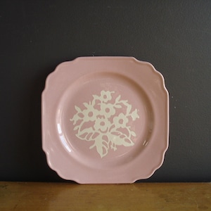 Pode incluir: Um prato quadrado de cerâmica rosa com borda ondulada. O prato apresenta um desenho floral branco no centro. O prato é exibido em uma superfície de madeira contra um fundo cinza escuro. Uma peça decorativa para a casa.