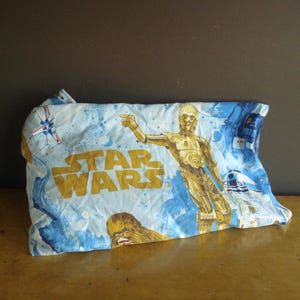 Puede incluir: Una funda de almohada vintage con temática de Star Wars. Presenta a C-3PO, R2-D2 y Chewbacca sobre un fondo azul claro, con las palabras "STAR WARS" en dorado. El diseño es retro.