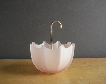Puede incluir: Un plato de vidrio rosa en forma de paraguas con un mango de metal dorado. El plato está sobre una superficie de madera.
