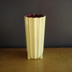 May include: A tall, cream-colored vase with a fluted design. The interior rim is a deep burgundy color. The vase is approximately 10 inches tall and has a rectangular base. The vase is made of ceramic or porcelain.