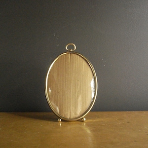 May include: An oval-shaped picture frame with a polished gold-tone metal finish. The frame features a small circular hanging loop at the top and two small spherical feet at the base, with a clear glass front covering a tan cardboard backing.