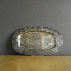 May include: A tarnished silver-coloured oval tray. The tray has a slightly raised rim and a rectangular interior. The surface shows signs of wear and age. The tray is set against a dark grey background and a wooden surface.