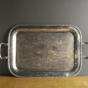 May include: A rectangular silver-toned metal serving tray with two curved handles on the sides. The tray features an ornate, embossed floral pattern along the border and a central area with a tarnished, weathered finish and intricate engravings.