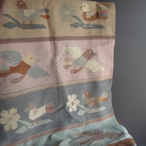 May include: Woven textile featuring a repeating pattern of stylized birds and flowers in muted tones. The design includes birds with outstretched wings, set against a backdrop of beige, pink, and blue-gray stripes. The textile has a textured appearance.