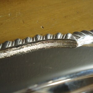 May include: Close-up of a silver-colored tray with a decorative, ridged edge. The tray's surface reflects light, and the edge has a detailed, textured design. The background is a light brown wooden surface.
