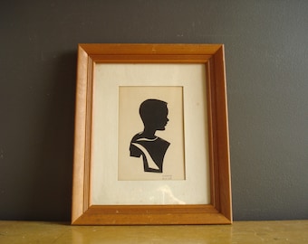 Könnte beinhalten: Eine gerahmte Silhouette eines Kopfes und der Schultern einer Person, aus schwarzem Papier ausgeschnitten und auf einen weißen Hintergrund montiert. Die Silhouette ist nach rechts gerichtet und die Person hat kurzes Haar. Der Rahmen ist aus Holz und hat eine hellbraune Oberfläche.