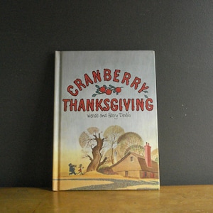 May include: A hardcover book titled Cranberry Thanksgiving by Wende and Harry Devlin. The cover features a light-colored background with red text, an illustration of three cranberries, a large tree, a house with a chimney, and two figures.