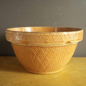 May include: A vintage, light brown mixing bowl with a decorative heart and triangle pattern. The bowl has a wide rim with a repeating heart design. The exterior features a textured pattern of hearts and triangles. The bowl is likely made of ceramic.
