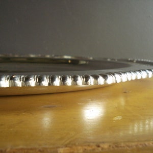 May include: A silver-colored, round serving tray with a beaded edge. The tray is set on a wooden surface, with a dark gray background. The tray's edge has a repeating pattern of small, rounded beads.
