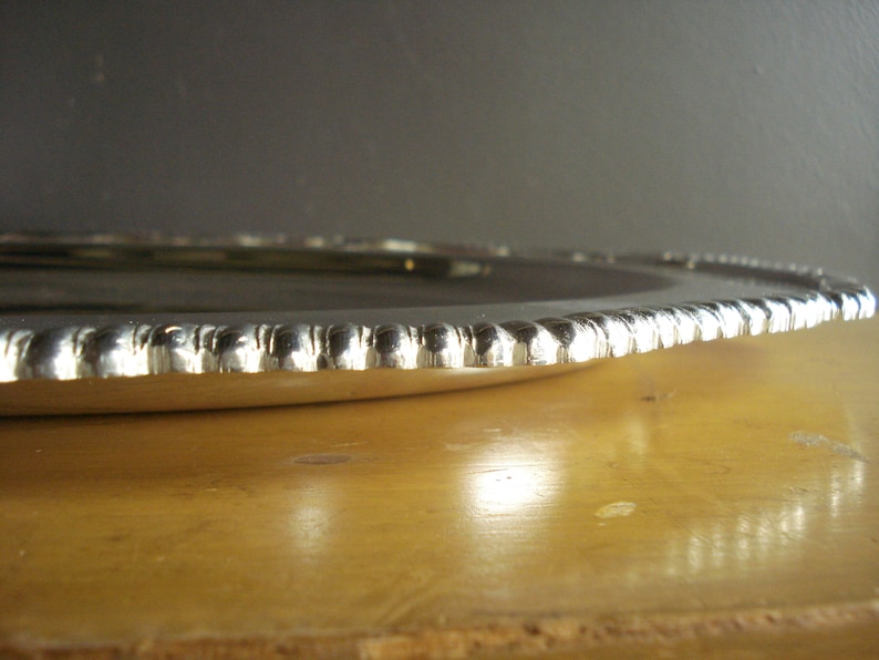May include: A silver-colored, round serving tray with a beaded edge. The tray is set on a wooden surface, with a dark gray background. The tray's edge has a repeating pattern of small, rounded beads.