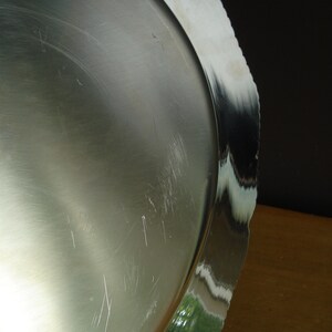 May include: A silver-colored, round serving tray with a scalloped edge. The tray has a reflective surface with visible scratches. The edge reflects the surrounding environment, showing green and white colors.