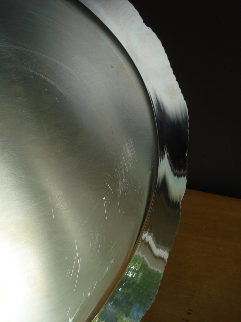 May include: A silver-colored, round serving tray with a scalloped edge. The tray has a reflective surface with visible scratches. The edge reflects the surrounding environment, showing green and white colors.