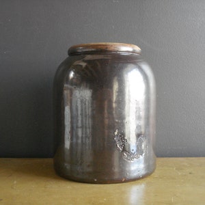 May include: A large, dark brown glazed stoneware crock with a wooden lid. The crock has a smooth, shiny surface and is slightly rounded at the bottom.