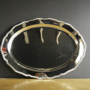 May include: Oval silver serving tray with a decorative rope-like edge. The tray has a highly reflective surface, and the rim has a scalloped design. The tray is displayed on a wooden surface against a dark background.