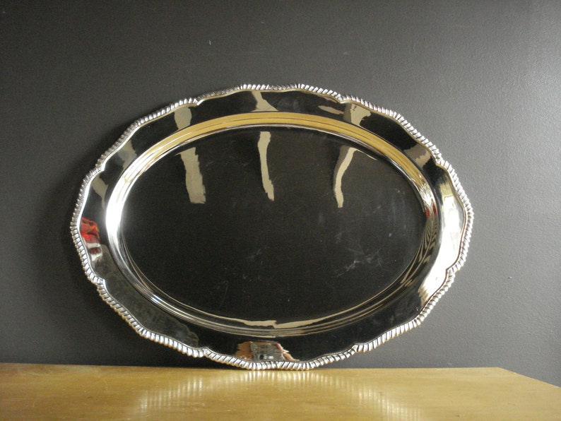 May include: Oval silver serving tray with a decorative rope-like edge. The tray has a highly reflective surface, and the rim has a scalloped design. The tray is displayed on a wooden surface against a dark background.