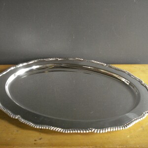 May include: Oval silver serving tray with a scalloped edge. The tray has a reflective surface and a decorative rope-like border. The tray is displayed on a light-colored wooden surface.