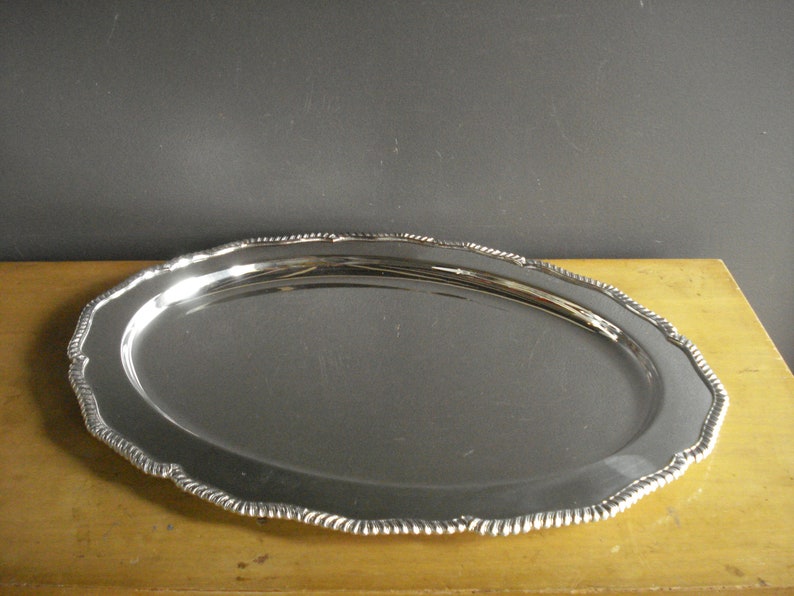 May include: Oval silver serving tray with a scalloped edge. The tray has a reflective surface and a decorative rope-like border. The tray is displayed on a light-colored wooden surface.