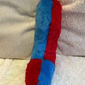 以下が含まれることがあります： 鮮やかな青と赤の、細長いぬいぐるみ。 ぬいぐるみは、青と赤の2つの縦のセクションに分かれており、ふわふわした質感です。 上部には黒いループが付いています。