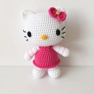 Peut inclure: Une poupée Hello Kitty en crochet blanc portant une robe rose avec une fleur rose sur la tête. La poupée a des yeux noirs et un nez jaune.