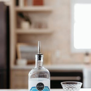 May include: A clear glass bottle with a metal pour spout, labelled "SIP N SADDLE BLUEBERRY LEMON VODKA SPRITZ." A cut crystal cocktail glass and a bar tool are on the white countertop. The background is blurred.