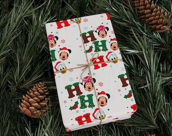 Puede incluir: Un regalo envuelto con temática navideña. El papel de regalo blanco presenta a Mickey y Minnie Mouse, Donald Duck y la frase "HO HO HO". El regalo está atado con hilo y colocado sobre un fondo de agujas de pino y piñas.