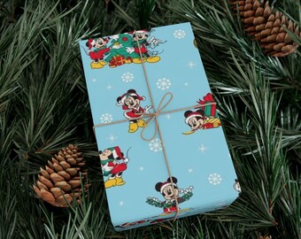 Puede incluir: Un regalo envuelto con un fondo azul claro que presenta un patrón repetido de Mickey Mouse con atuendo navideño y copos de nieve. El regalo está atado con hilo y colocado sobre un fondo de agujas de pino y piñas.