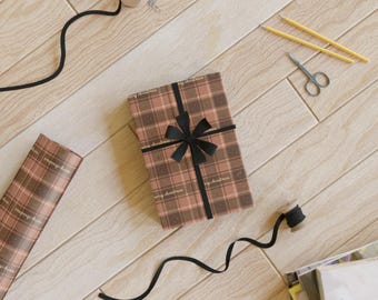 Puede incluir: Un regalo envuelto con un lazo y cinta negros, papel de regalo, tijeras y lápices, dispuestos sobre una superficie de madera clara. El papel de regalo tiene un patrón de cuadros y las palabras "merry christmas".