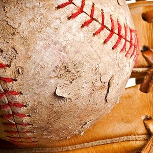 Op de afbeelding: Close-up van een versleten honkbal met rode stiksels en een bruinleren honkbalhandschoen. De honkbal vertoont tekenen van slijtage, met krassen en verkleuring. De handschoen is gemaakt van bruin leer.