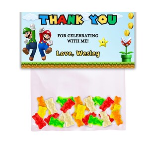 May include: A thank you favor bag with a Mario and Luigi themed design. The bag has a blue and green background with the text "Thank You" in a colorful font. The bag also includes the text "For celebrating with me! Love, Wesley". The bag is filled with colorful gummy bears.