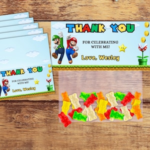 May include: Thank you favor bags with a blue background and a cartoon image of Mario and Luigi. The bags are filled with colorful gummy candies. The text on the bag reads "Thank You for celebrating with me! Love, Wesley".