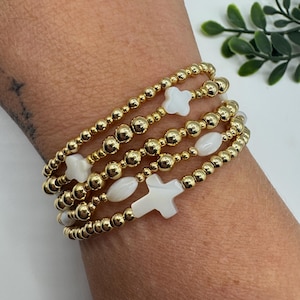 May include: Gold beaded bracelets with white clover, cross, and oval-shaped accents. The bracelets are stacked on a wrist, showcasing the jewelry's design and texture. The gold beads and white accents create a contrast, highlighting the details of each bracelet.