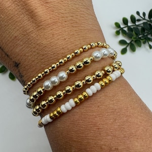 May include: A collection of gold beaded bracelets with pearl and white bead accents. The bracelets are stacked on a wrist, showcasing the different textures and designs. The gold beads vary in size, creating a layered look.