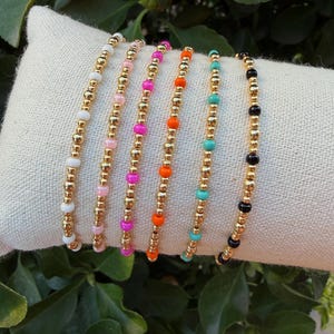May include: Six beaded bracelets arranged in a row, each featuring a repeating pattern of metallic gold beads interspersed with single colored beads in white, light pink, hot pink, bright orange, turquoise, and black, creating a vibrant set.