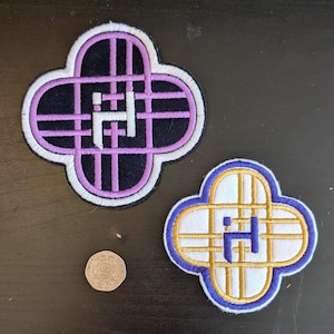 Könnte beinhalten: Zwei gestickte Aufnäher in Kleeblattform. Einer hat einen schwarzen Hintergrund mit lila und weißen Details, der andere einen weißen Hintergrund mit goldenen und blauen Details. Eine Münze ist ebenfalls vorhanden.