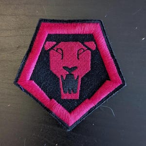 May include: Embroidered patch with a red lion's head on a black background, enclosed by a red border. The patch is pentagon-shaped. The lion's mouth is open, and the design is detailed with visible stitching.