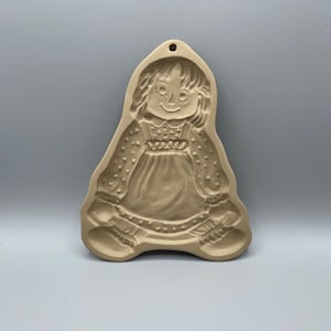 Könnte beinhalten: Ein beiger Keramik-Ausstecher in Form einer Puppenfigur mit einem einfachen Kleid und einem lächelnden Gesicht.