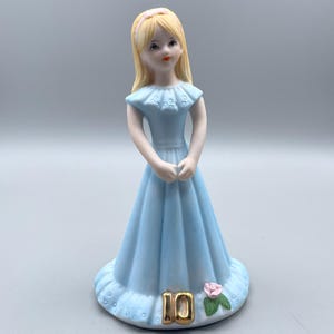 Pode incluir: Uma figurinha de porcelana de uma menina com um vestido azul claro e uma flor rosa na base. O número 10 está escrito em ouro na base.