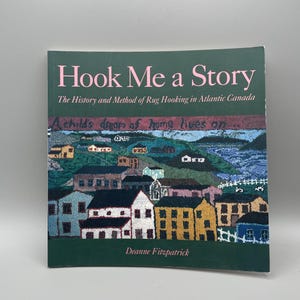 May include: A softcover book titled Hook Me a Story: The History and Method of Rug Hooking in Atlantic Canada by Deanne Fitzpatrick. The cover features a colorful, textured rug-hooked illustration of a coastal village with houses and water.