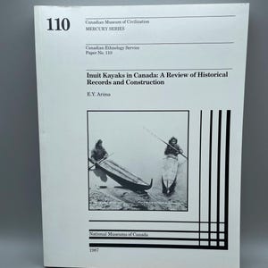 May include: A book cover titled "Inuit Kayaks in Canada: A Review of Historical Records and Construction" by E.Y. Arima. The cover shows a black and white photograph of two people in kayaks. Part of the Mercury Series, published by the Canadian Ethnology Service.