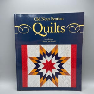 Może przedstawiać: Książka zatytułowana "Old Nova Scotian Quilts" z granatową okładką i złotymi literami. Okładka przedstawia wzór pikowania z czerwonym, białym, granatowym i pomarańczowym wzorem gwiazdy. Poniżej tytułu znajdują się nazwiska autorów, Scotta Robsona i Sharon MacDonald.