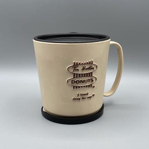 Puede incluir: Una taza de cerámica beige con tapa y asa negras. La taza presenta el logotipo de Tim Hortons con el texto "Tim Hortons Donuts. A friend along the way."