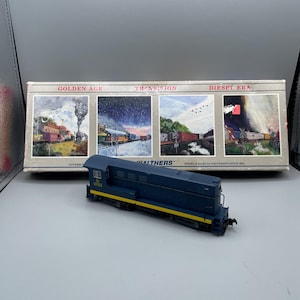 Puede incluir: Una locomotora de tren modelo azul y amarilla con el número 5703 en el lateral. La locomotora está sobre una superficie blanca delante de una caja con las palabras "Golden Age", "Transition" y "Diesel Era" en ella. La caja también tiene imágenes de trenes en diferentes entornos.